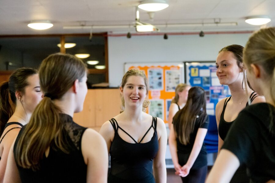 Teenage girl with blonde hair speaks to group of other teenage girls wearing dance clothes. All are smiling.